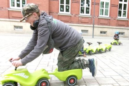 Ein Jugendlicher, seitlich zu sehen, kniet auf einem grünen Bobby-Car mit Anhänger. Die Knie auf dem Sitz, beide Arme greifen das Lenkrad. Im Hintergrund vier weitere Bobby-Cars und ein Kind.